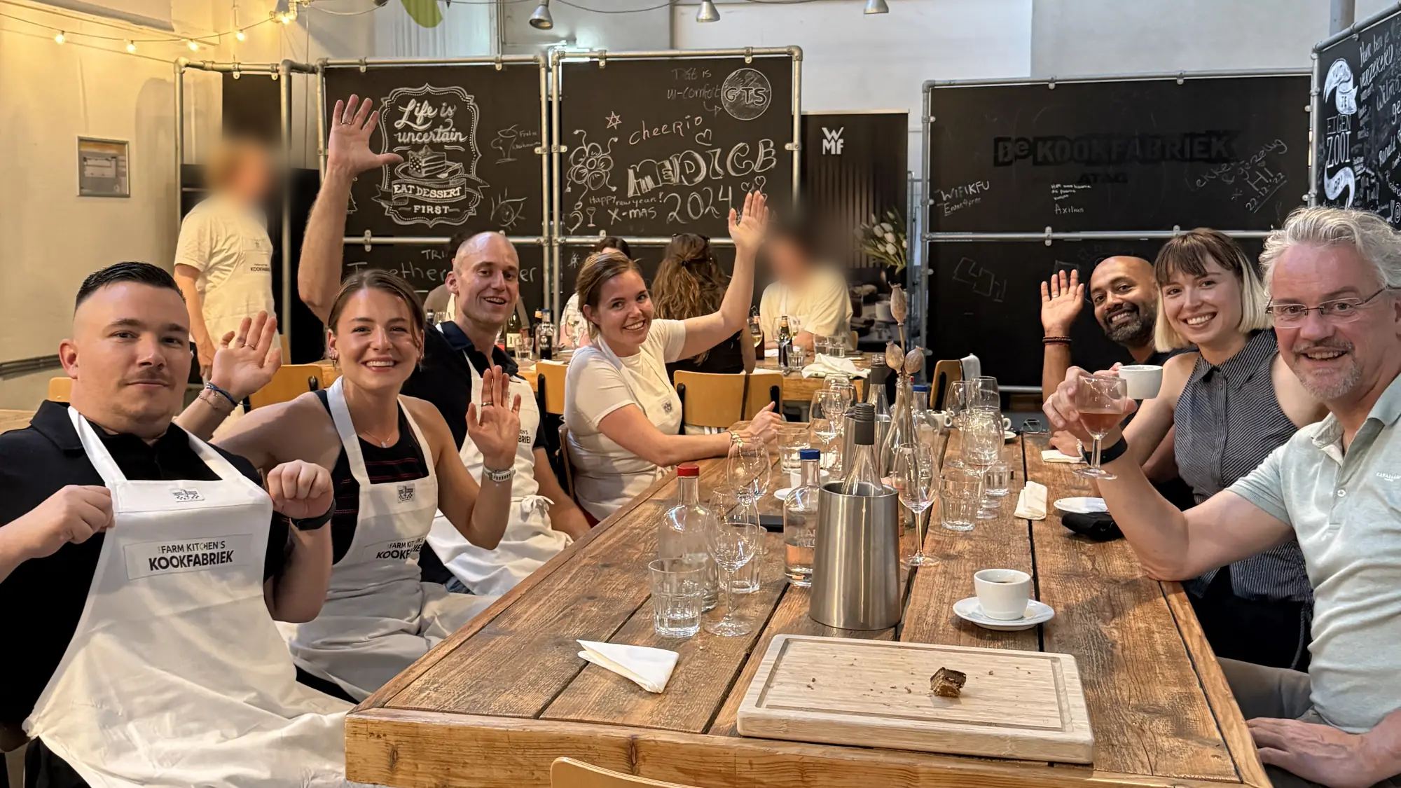 Het Axveco-team aan een lange tafel in een kookstudio, een aantal met de hand omhoog.